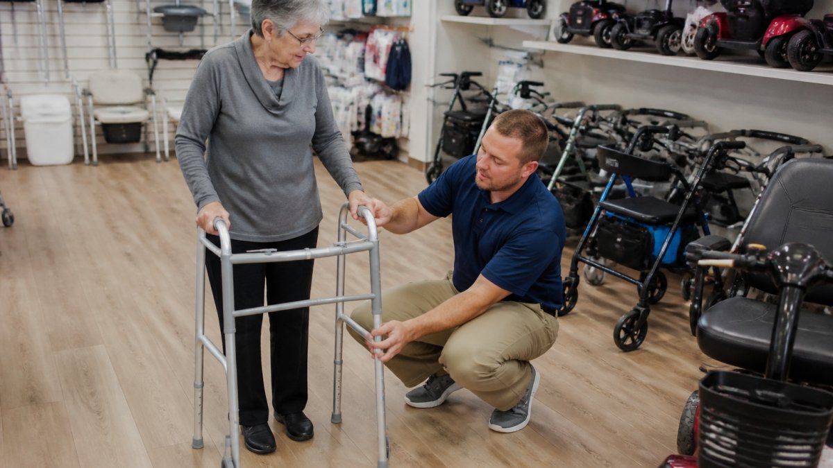 Senior adult being fitted with a mobility walker by a knowledgeable medical equipment specialist in a bright showroom with various mobility aids displayed on shelves