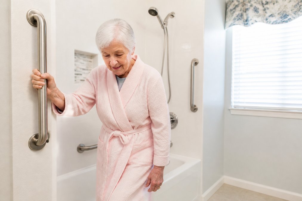 A senior woman gripping a securely mounted stainless steel grab bar next to a bathtub in a bright bathroom, showing proper placement and confident use