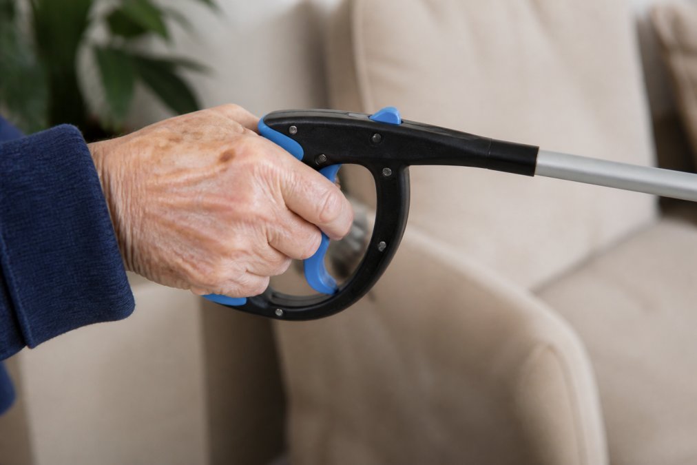 Close-up of a person's hand with visible joint changes holding a reacher tool with an ergonomic handle, demonstrating proper grip position and trigger mechanism engagement in natural lighting